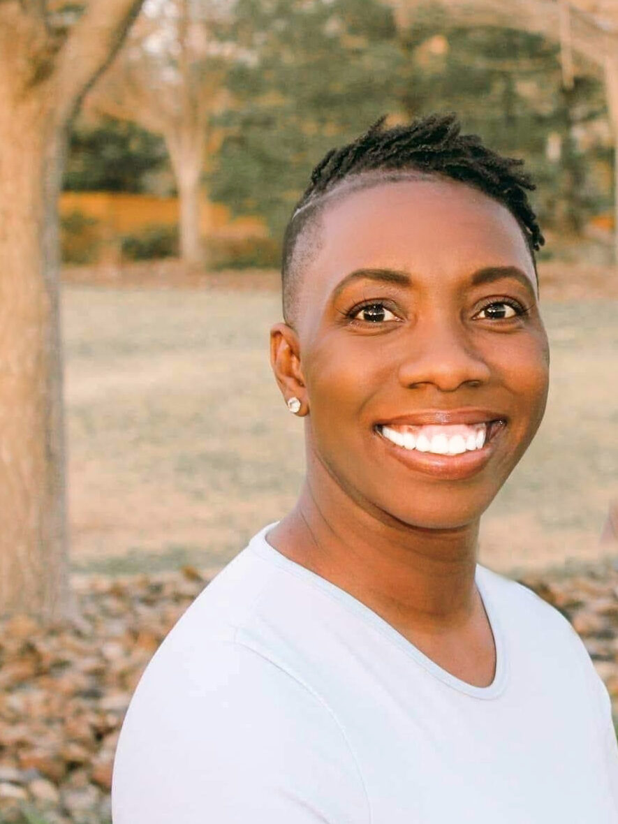 Me: A dark skin black woman poses with a smile in front of leafy trees