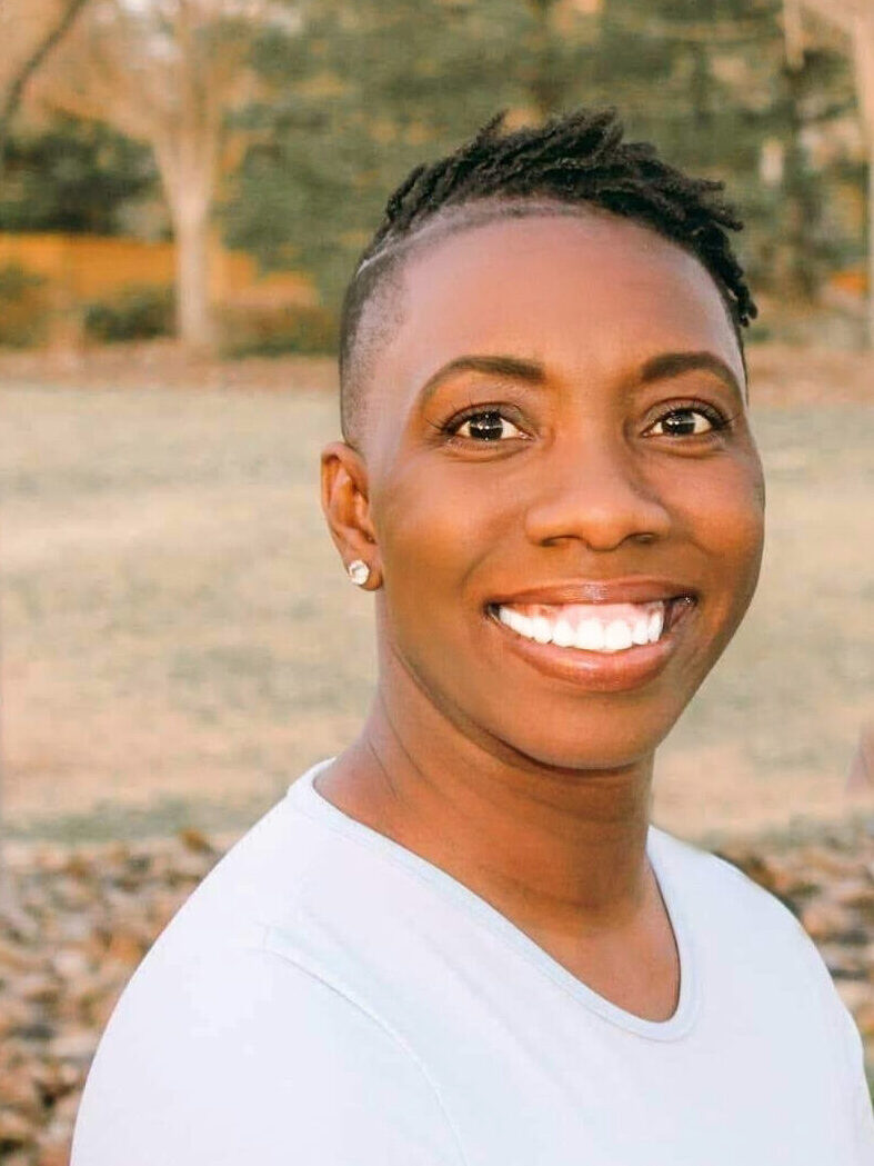 Me: A dark skin black woman poses with a smile in front of leafy trees